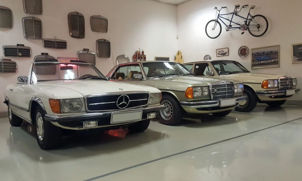 A R107 SL next to a C123 (W123 coupe) variant and W123, on the wall behind are numerous old Mercedes front grilles.